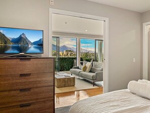 A bedroom with a bed, a dresser, a television, and a view of a mountainous landscape through a sliding door. - Newly Renovated Lakefront Retreat, Sleeps 2 (Queenstown)