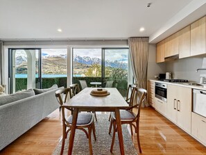 A modern kitchen with wooden cabinets, a dining area with a table and chairs, and a large window offering a view of mountains and a lake. - Newly Renovated Lakefront Retreat, Sleeps 2 (Queenstown)