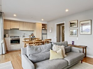 A modern living room with a grey sofa, wooden dining table, and kitchen in the background. - Newly Renovated Lakefront Retreat, Sleeps 2 (Queenstown)
