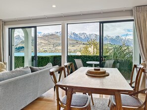 A dining area with a wooden table and chairs, a grey sofa, and large windows offering a mountain view. - Newly Renovated Lakefront Retreat, Sleeps 2 (Queenstown)