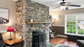 Una sala de estar con chimenea de piedra, ventilador de techo, ventana con persianas y estantería.