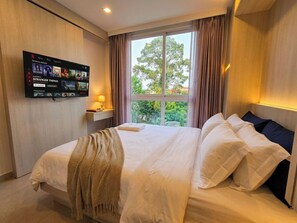 Habitación de hotel con una cama, un televisor montado en la pared, una ventana con cortinas y una mesita de noche con una lámpara.