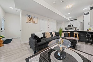 Un salon moderne avec un canapé gris, une table basse en verre et une cuisine en arrière-plan.