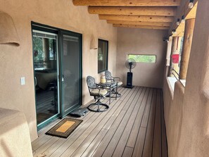 A covered patio with a wooden floor, a dining table with chairs, a standing fan, and a sliding glass door leading to an outdoor area. - Unique 1-bedroom Adobe casa apartment with beautiful deck in Albuquerque, (Albuquerque)