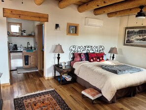 Ein Schlafzimmer mit Bett, Nachttischlampen, einem Nachttisch, einem kleinen Hocker, Holzboden, einer Küchenzeile mit Kühlschrank und einem Fenster.