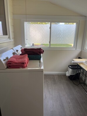 A laundry area with a washing machine, stacked towels, a window with frosted glass, and a wooden floor. - Cozy 2-bedroom cottage in a country setting in Ruffin NC (Ruffin)