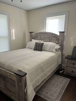 A bedroom with a wooden bed frame, a matching nightstand, a patterned rug, and a window with blinds. - Cozy 2-bedroom cottage in a country setting in Ruffin NC (Ruffin)