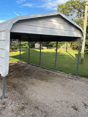A covered outdoor area with a gravel floor, metal supports, and a view of a grassy yard with a shed and trees. - Cozy 2-bedroom cottage in a country setting in Ruffin NC (Ruffin)