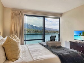 A hotel room with a bed, a chair, a television, and a view of a body of water and mountains. - Alpine Lakefront Villa, 2 Bath, Queenstown (Queenstown)