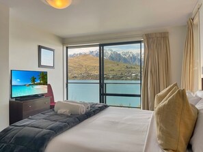 A hotel room with a bed, a television, and a large window offering a mountain and lake view. - Lakefront Villa, Sleeps 6, Walk to Queenstown Trail (Queenstown)