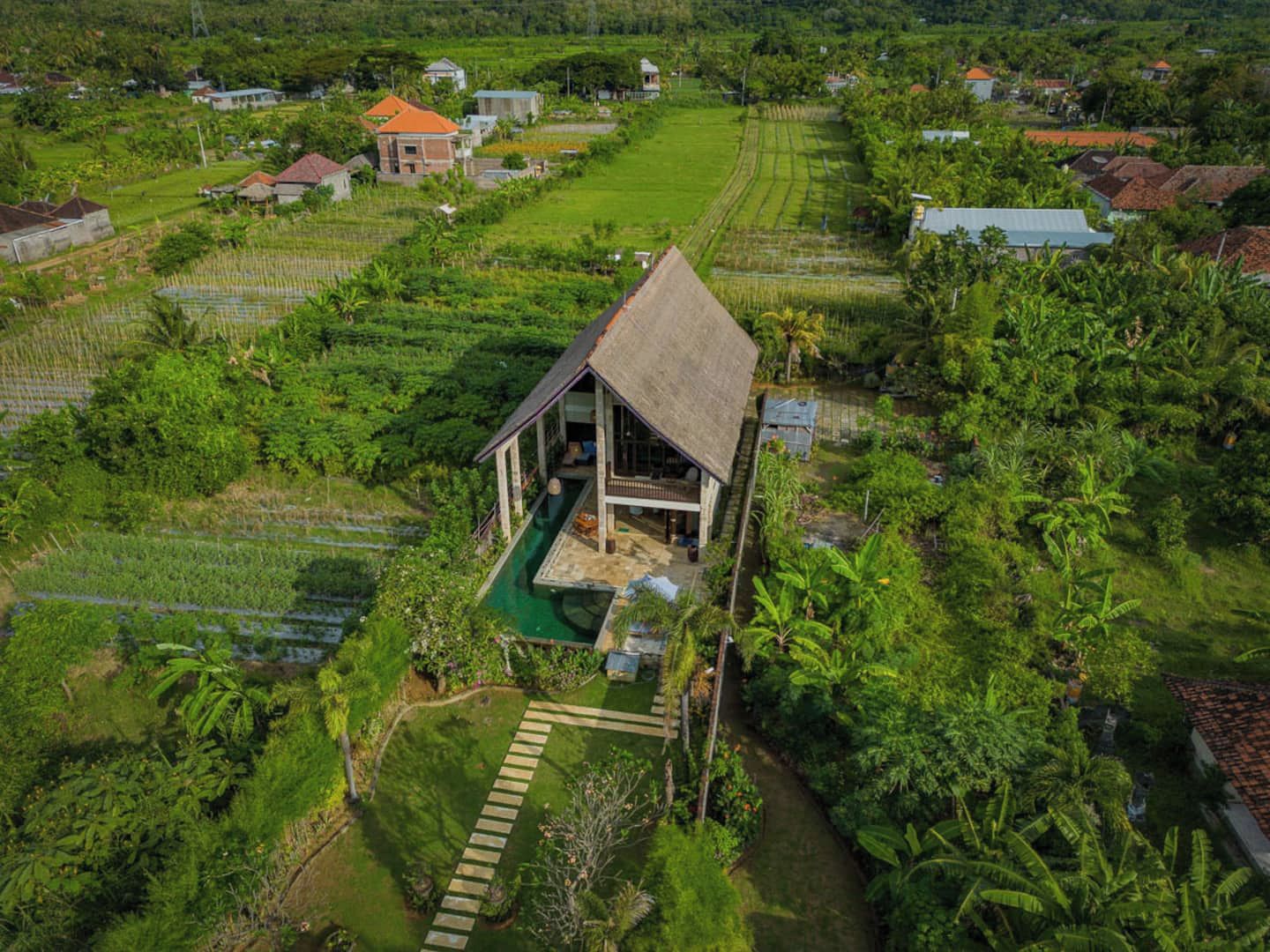 A single-story house with a thatched roof, surrounded by lush greenery and agricultural fields.