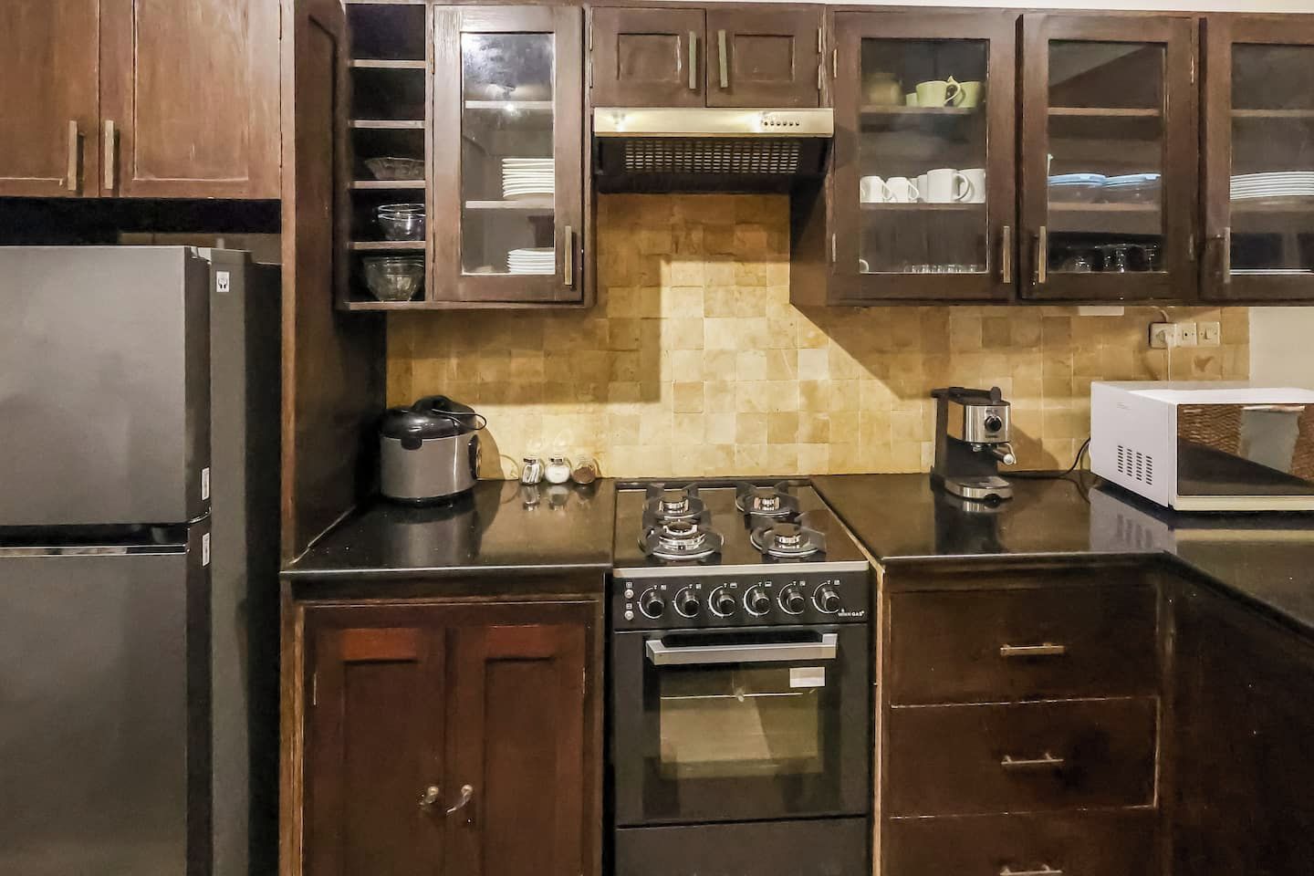 Una cocina con un refrigerador de acero inoxidable, una estufa con cuatro hornillas y un microondas. Hay armarios de madera con puertas de vidrio y un salpicadero de azulejos.