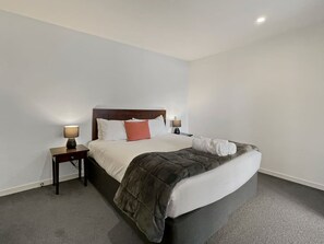 Quarto de hotel com uma cama de solteiro, mesa de cabeceira com abajur e uma parede branca lisa.