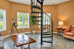 Une pièce avec un escalier en colimaçon, un télescope, une plante en pot, une table et un fauteuil à motifs.