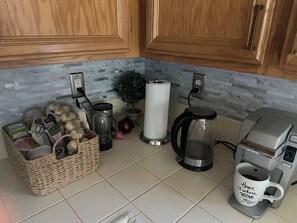Une cuisine avec des meubles en bois, une machine à café, une bouilloire, un panier d’épicerie et une plante en pot.