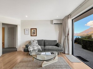 A modern living room with a grey sofa, a glass coffee table, and a view of a mountainous landscape through a sliding glass door. - Tranquil 1 Bed Corner Suite, Lake & Mountain Views (Queenstown)