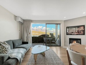 A modern living room with a sofa, coffee table, and dining table. There is a fireplace and a large window with a mountain view. - Tranquil 1 Bed Corner Suite, Lake & Mountain Views (Queenstown)