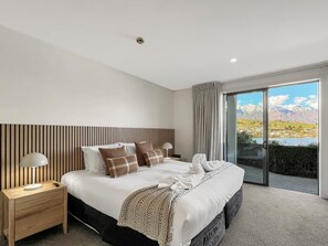 Ein modernes Schlafzimmer mit einem großen Bett, einem Holzkopfteil, einem Nachttisch, einer Lampe und Bergblick durch eine Schiebeglastür.