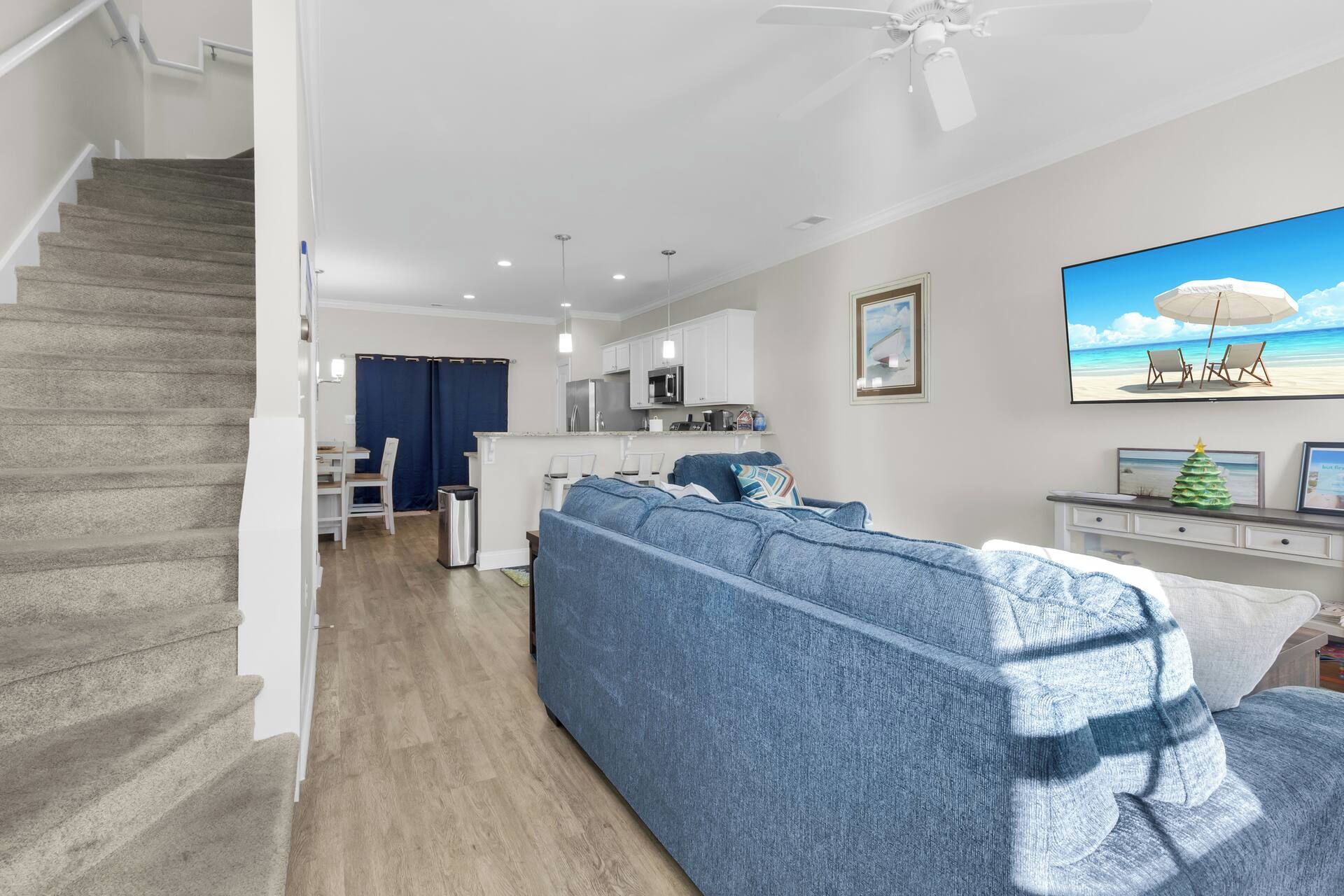 Ein modernes Wohnzimmer mit Treppe, einem blauen Sofa, einer Küchenzeile und einem Strandbild.