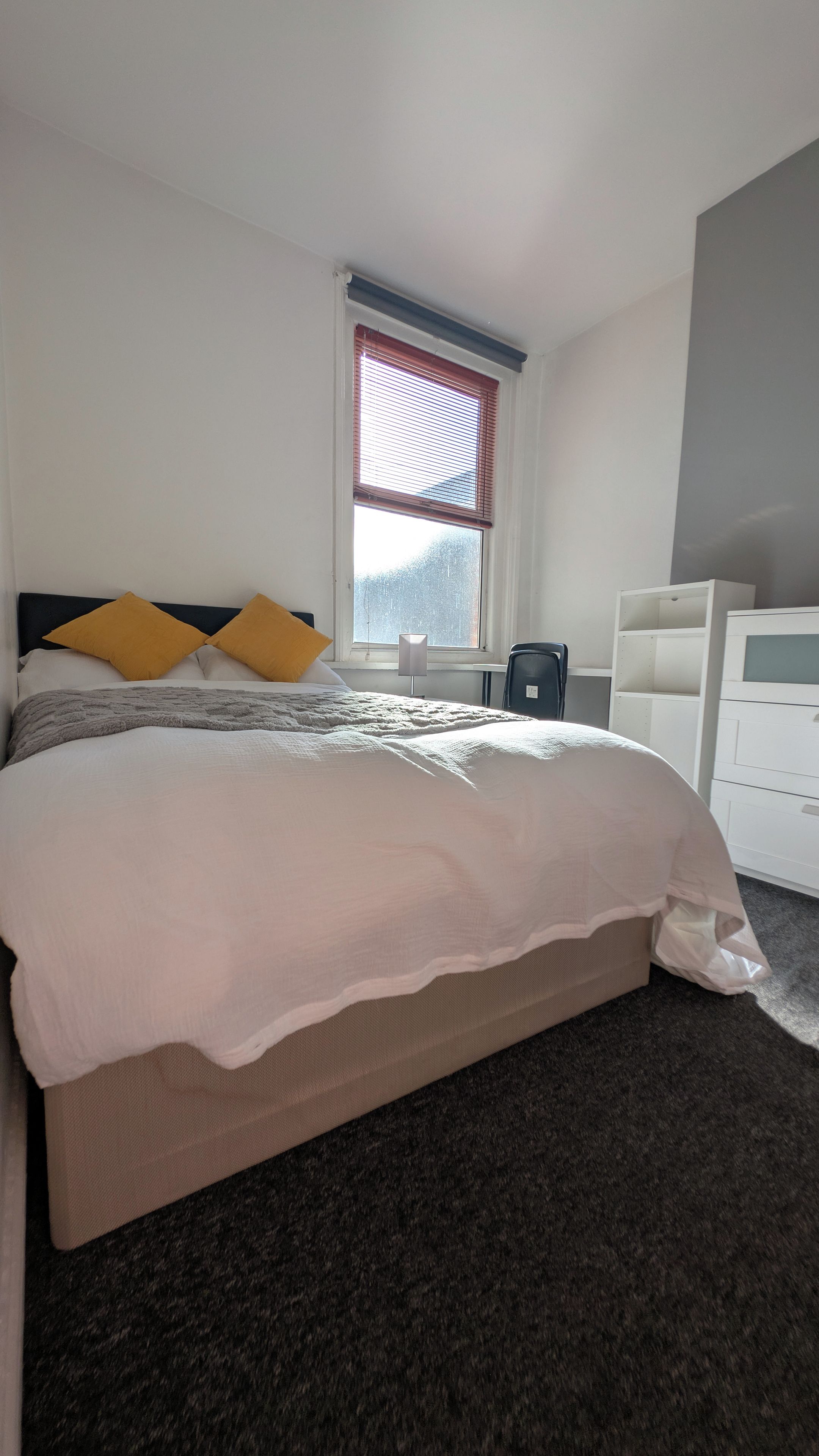 A bedroom with a bed, a window with blinds, a chair, and a white dresser.