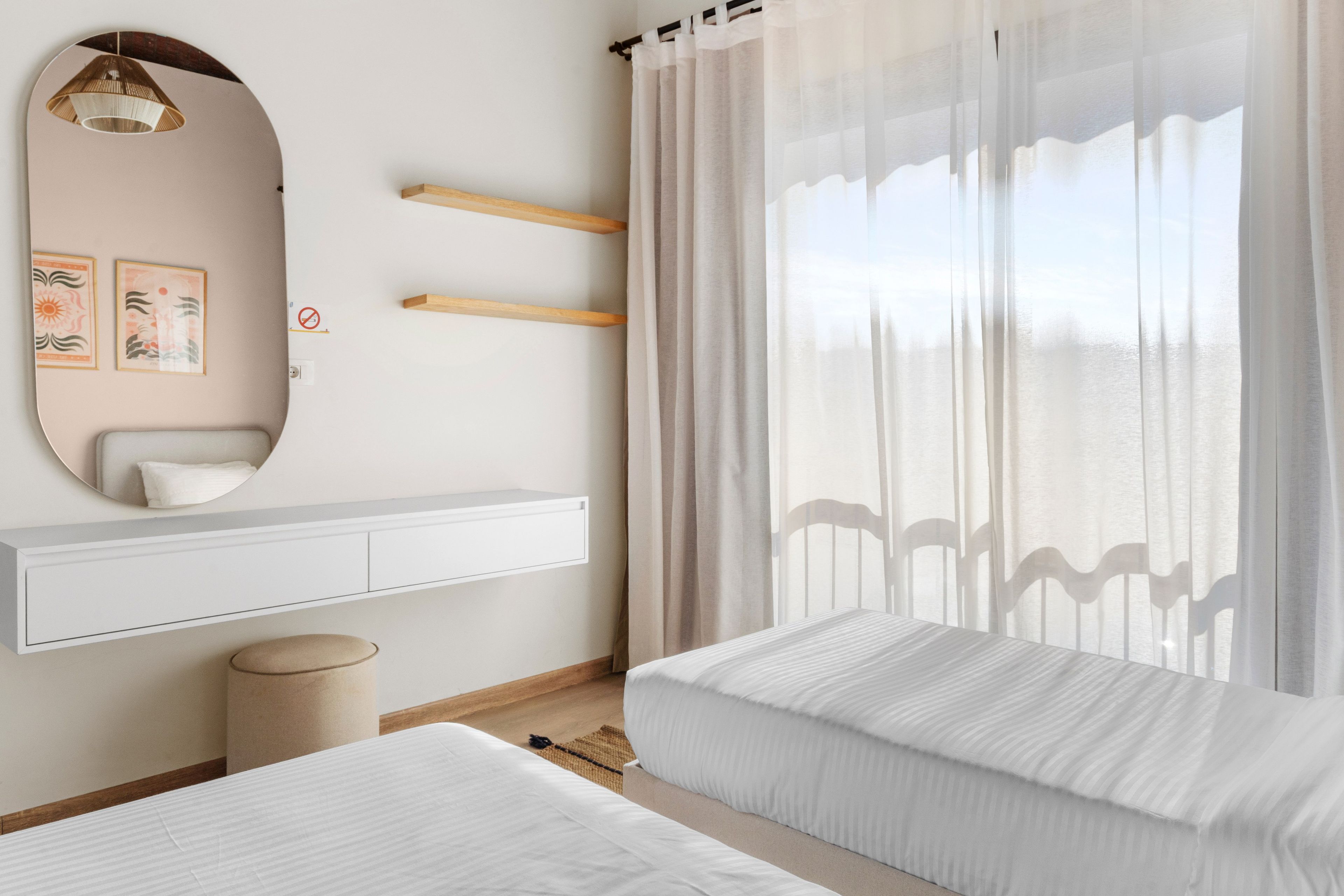 A bedroom with a bed, a bedside table, a mirror, and a window with sheer curtains.