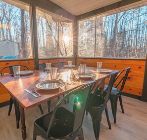 Comedor con mesa de madera dispuesta para cuatro, rodeada de sillas, y vista a árboles a través de amplias ventanas.