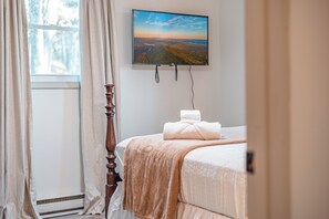 Ein Schlafzimmer mit Bett, einem Fenster mit Vorhängen, einem Bild an der Wand und einem Nachttisch mit Lampe.