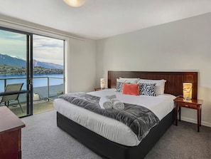 A bedroom with a large bed, a nightstand, a lamp, and a view of a lake and mountains. - Peaceful Alpine Villa, Lake Views, Sleeps 6 (Queenstown)
