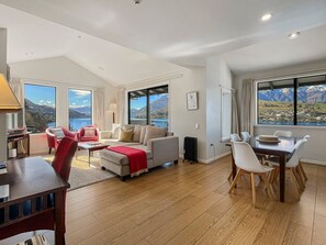 A spacious living area with a sofa set, a dining table with chairs, and large windows offering a view of mountains and a lake. - Peaceful Alpine Villa, Lake Views, Sleeps 6 (Queenstown)