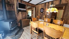 Ein rustikales Interieur mit Holzofen, einem Esstisch mit Stühlen, einem Fernseher und einer Wanduhr.