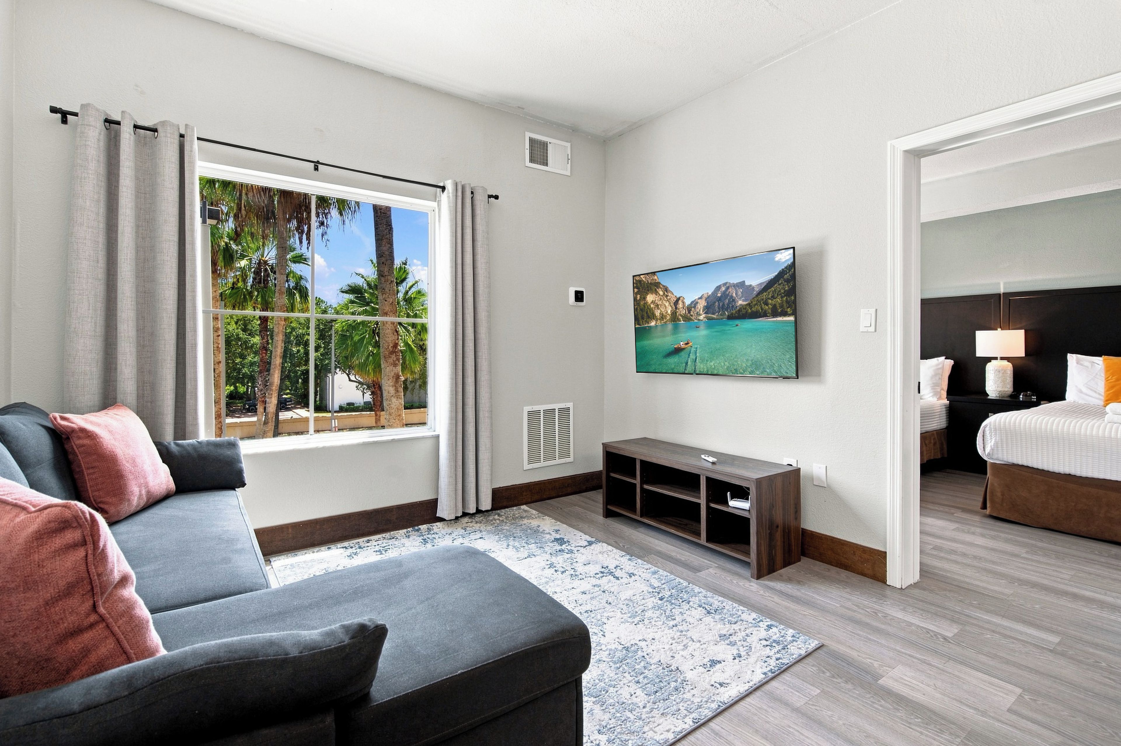 A hotel room with a grey sofa, a flat-screen TV mounted on the wall, a wooden floor, and a view of a mountainous landscape through the window.