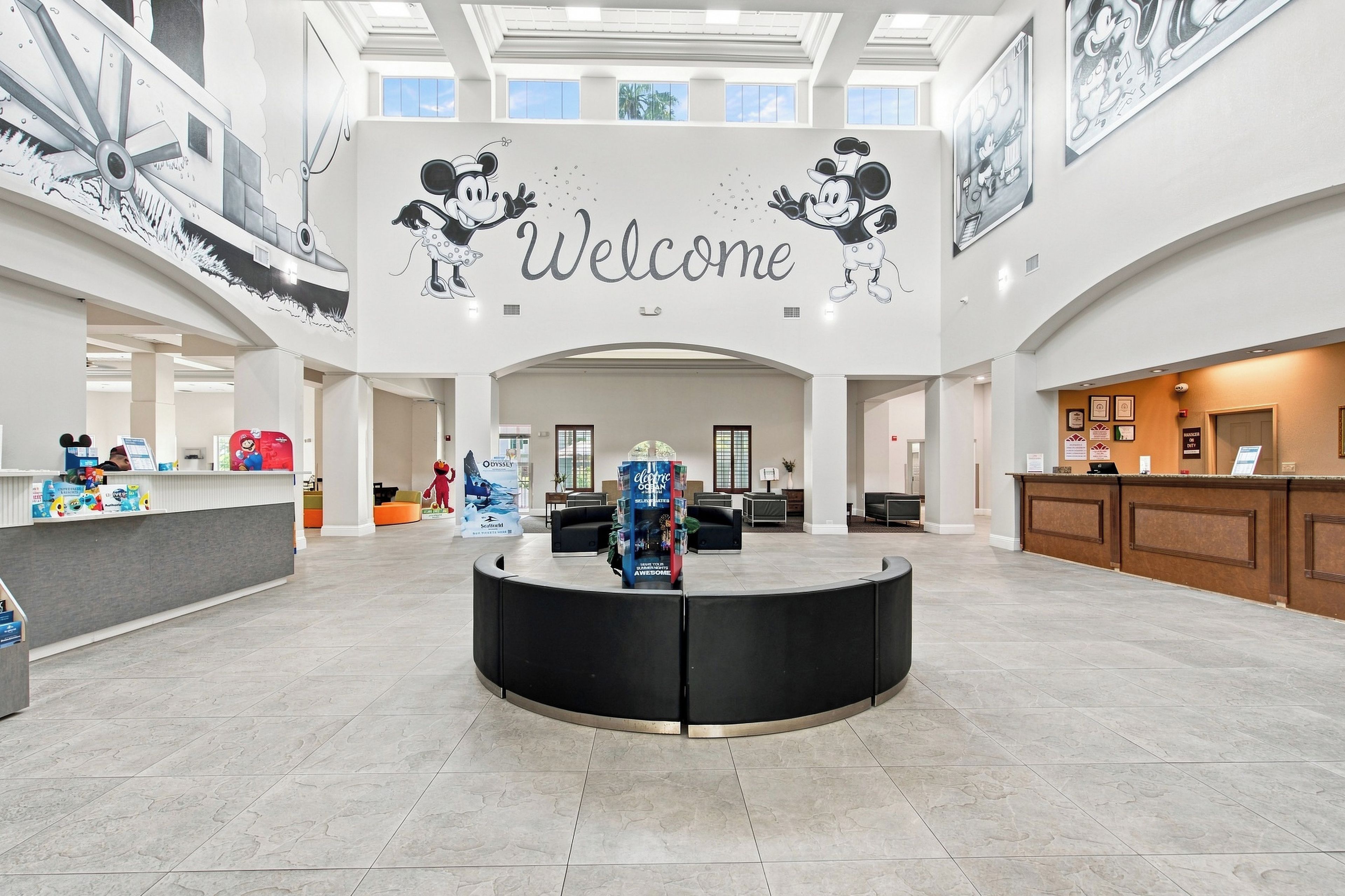 A spacious lobby with a central reception desk, a large welcome sign, and a high ceiling with skylights.