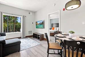 A modern living room with a sofa, a dining area with a table set for two, a television mounted on the wall, and a view of the outdoors through a window.