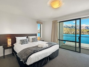 A hotel room with a large bed, a nightstand, a lamp, and a view of mountains and water. - Alpine Lakeview Villa, 3 Bed, Queenstown (Queenstown)
