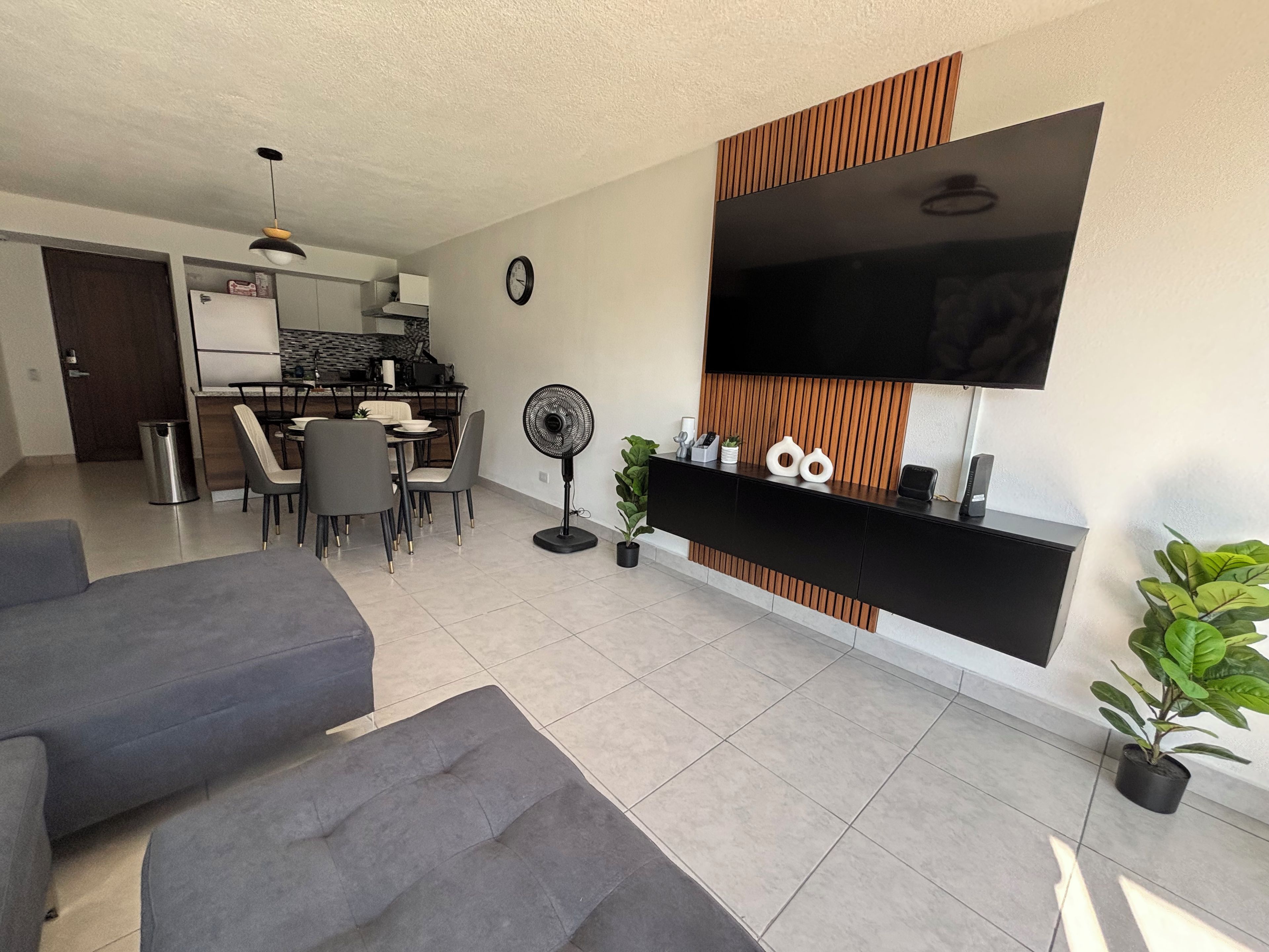Ein modernes Wohnzimmer mit einem Flachbildfernseher, grauen Sofas, einem Essbereich mit Stühlen und einer Küche im Hintergrund.