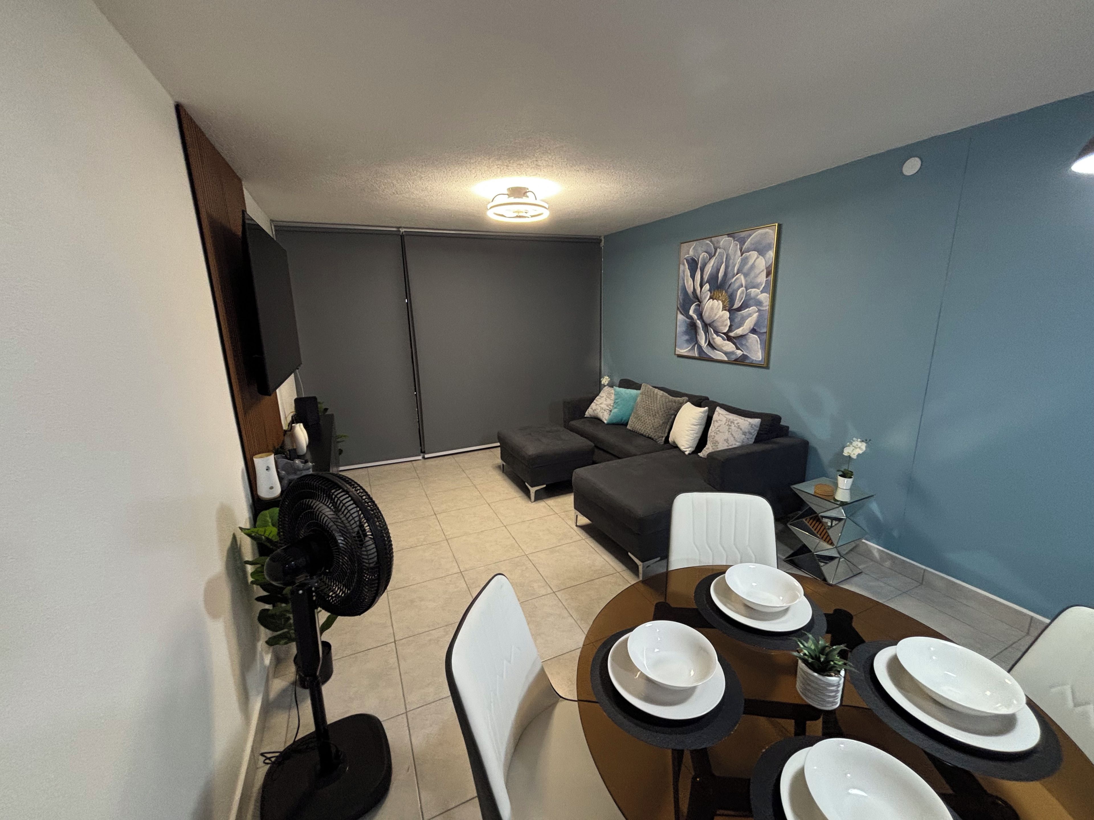 Una sala moderna con un sofá modular, una mesa de comedor preparada para cuatro personas y un ventilador de techo.