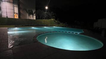 A nighttime swimming pool with a curved edge and a surrounding paved area.