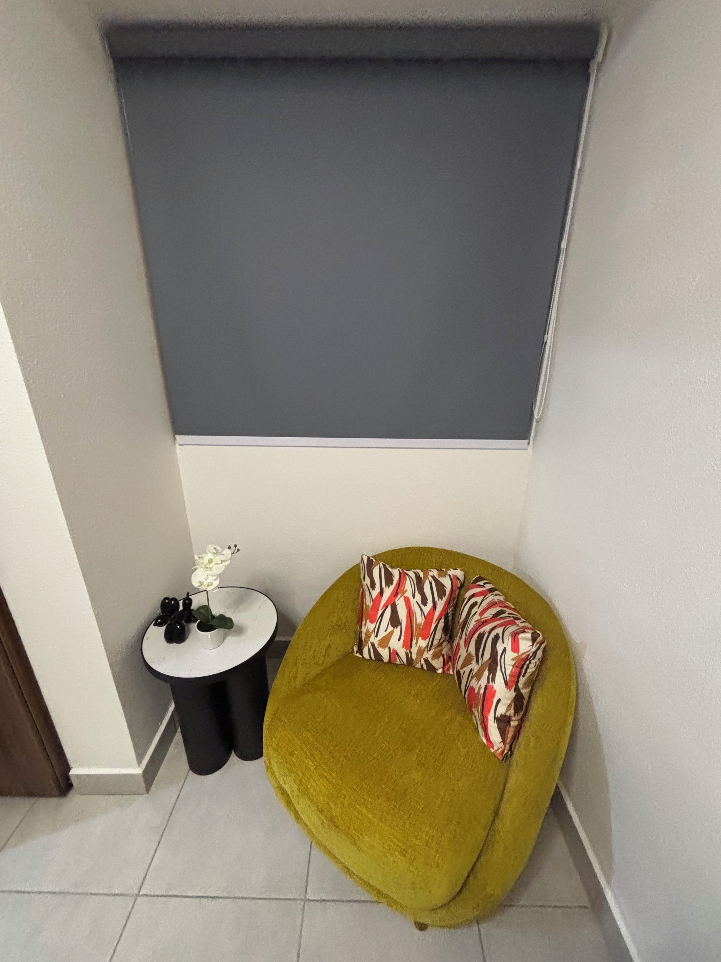 Una habitación de esquina con un sofá verde, una mesita redonda con un jarrón blanco y objetos negros, y una pared gris con persianas.