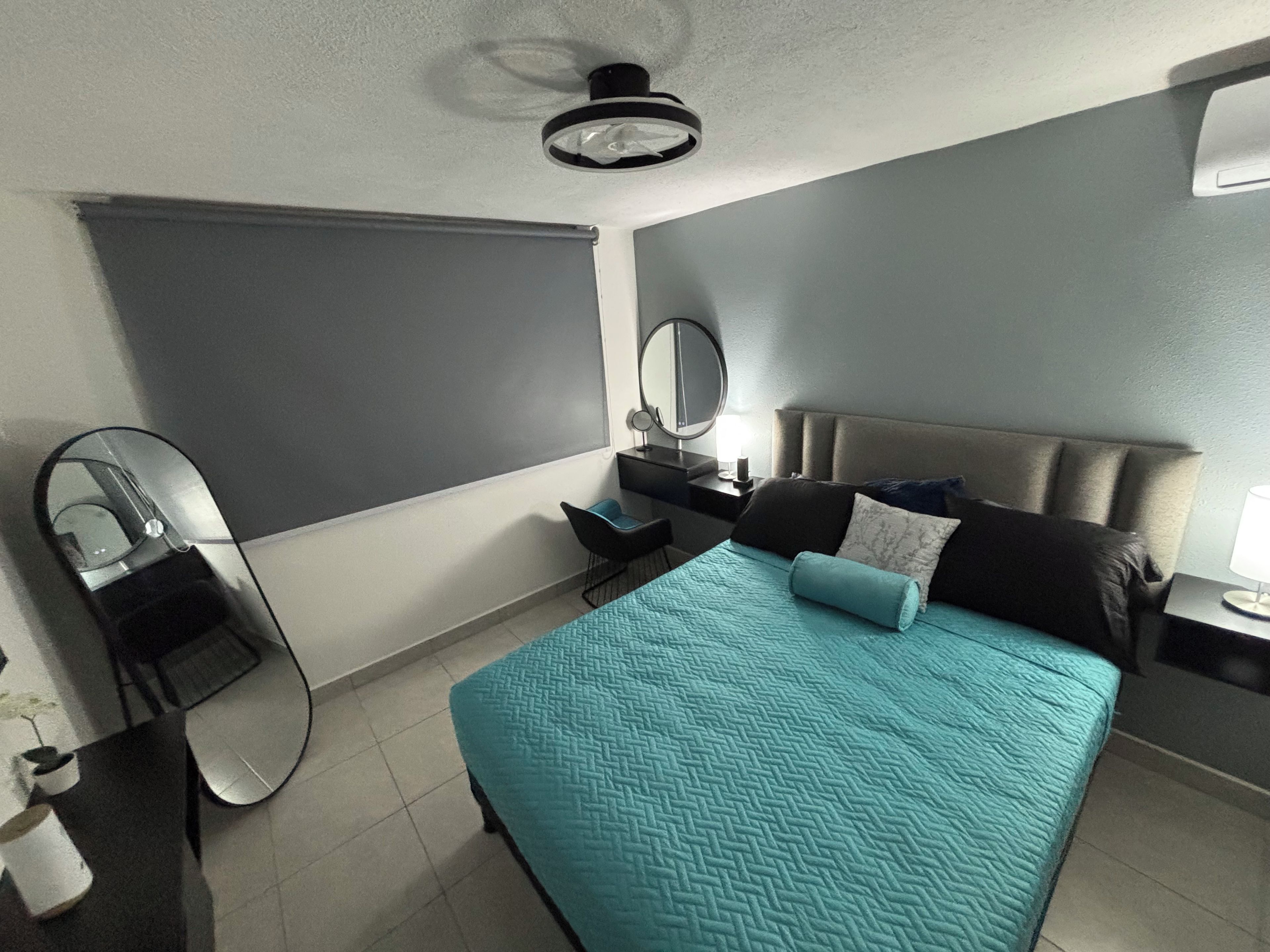 Une chambre d’hôtel comprenant un lit, une table de chevet, un miroir, une fenêtre avec un store et un ventilateur de plafond.