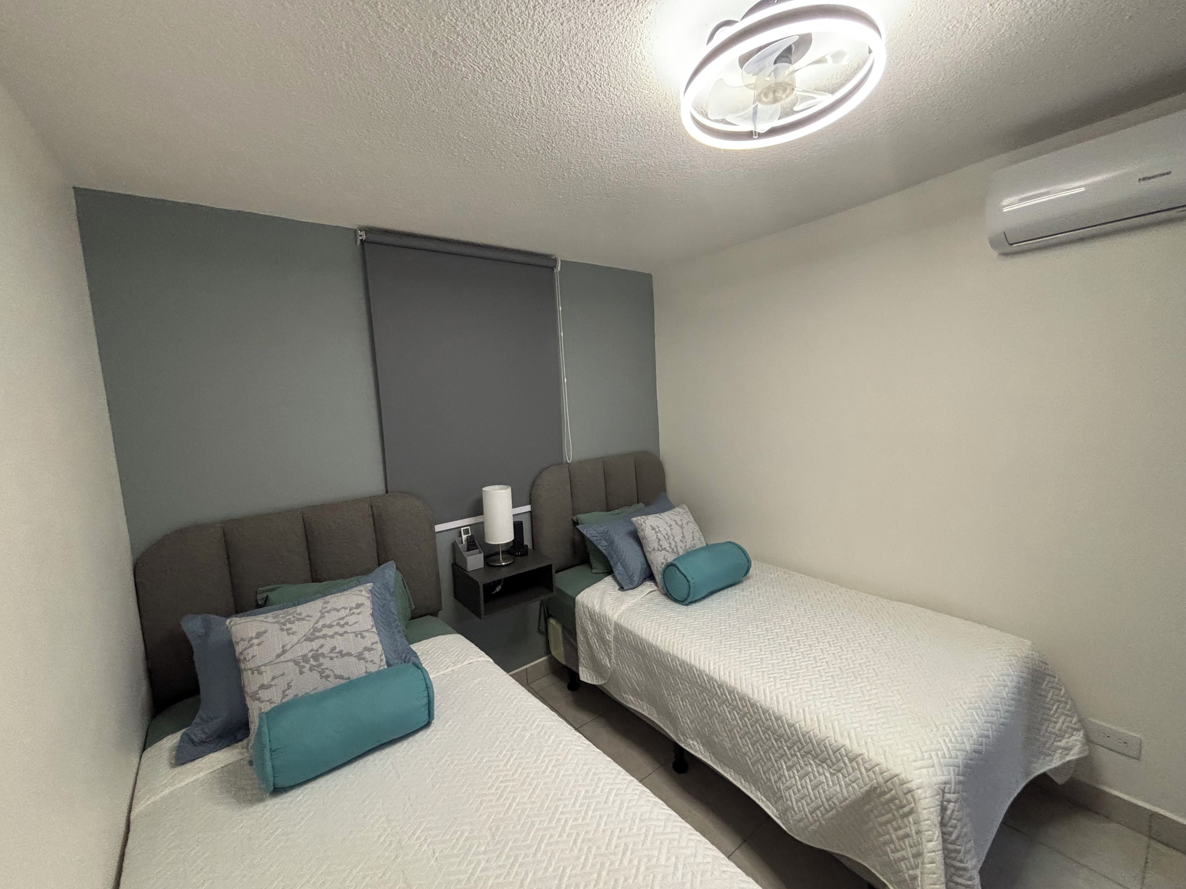 Une petite chambre à coucher moderne, avec deux lits simples, une table de chevet avec une lampe et un ventilateur de plafond.