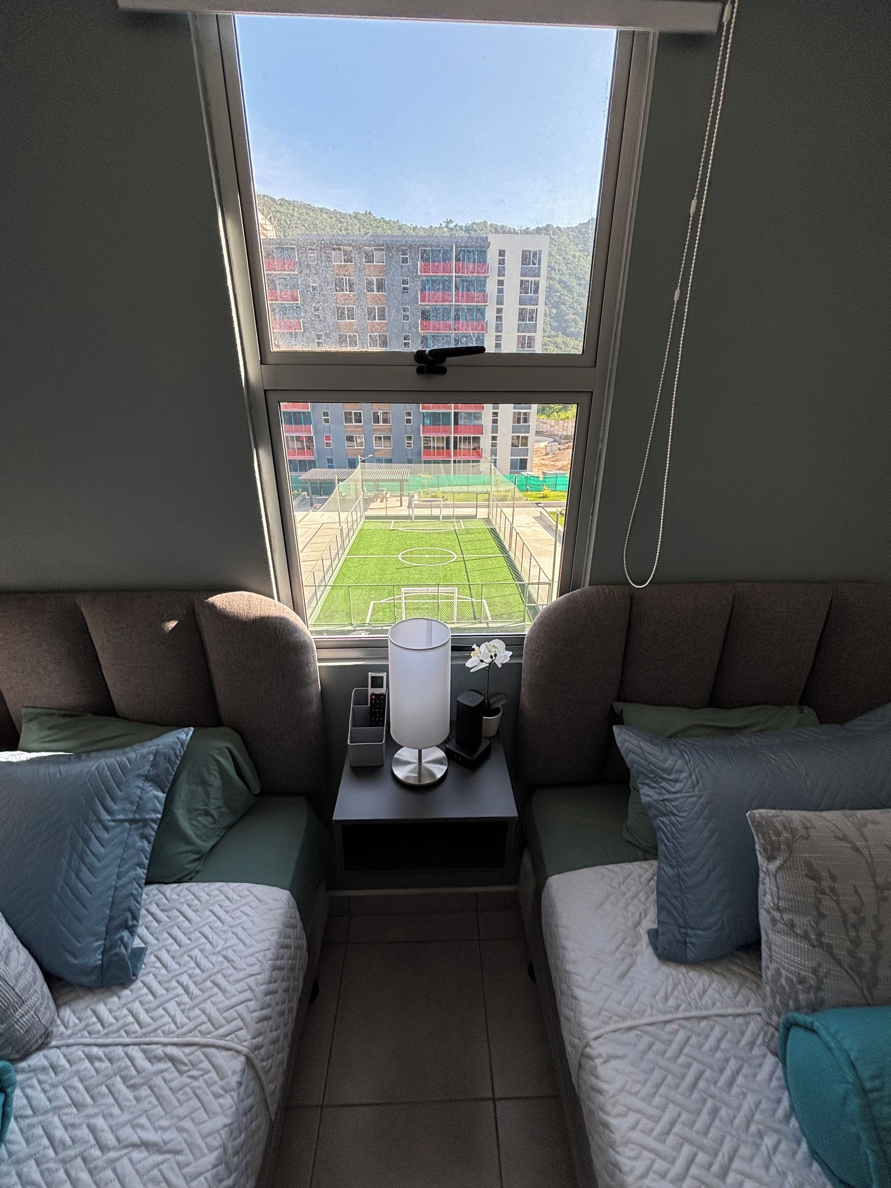 Una habitación con dos camas, una mesita con una lámpara, un teléfono y vistas a un edificio y un campo deportivo a través de una ventana.