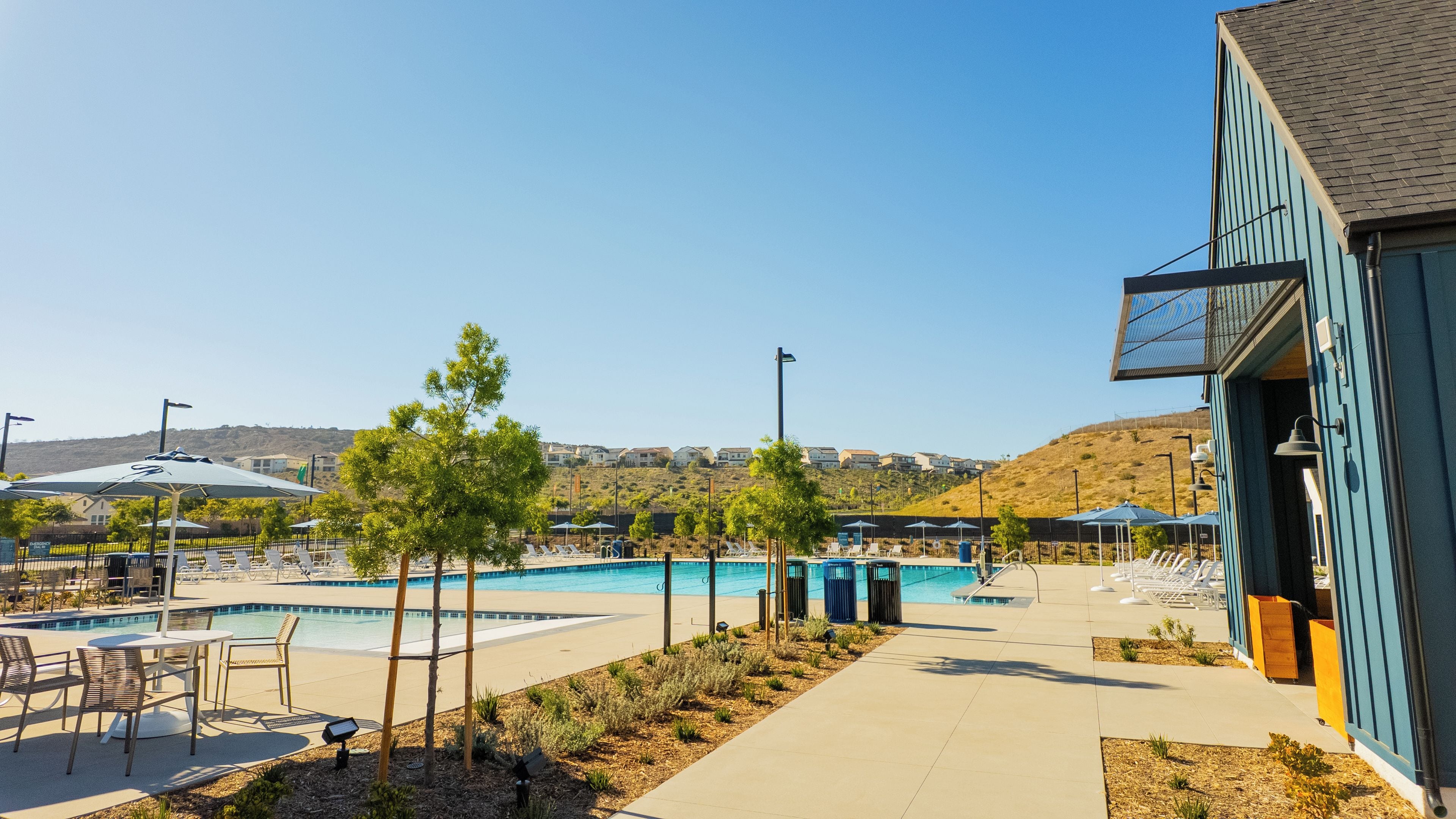 A pool area with lounge chairs and umbrellas, a clear sky, and a hillside in the background.