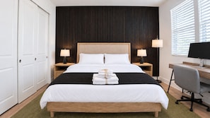 A hotel room with a large bed, two bedside tables with lamps, a desk with a computer, and a window with blinds.