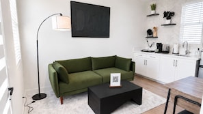 A modern living room with a green sofa, a black coffee table, a floor lamp, and a kitchen area with white cabinets and a countertop.