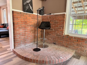 A corner room with a brick wall, a black chair, a microphone stand, and a window with a view.