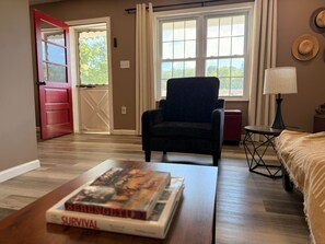 A room with a wooden floor, a black armchair, a small table with a book, a lamp, and a bed with a blanket.