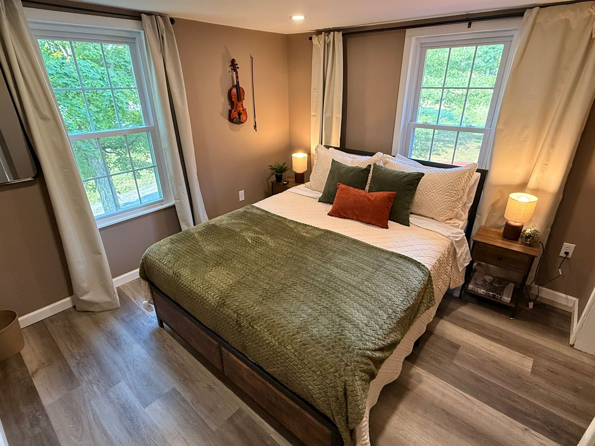 A bedroom with a bed, a violin on the wall, and two windows with curtains.