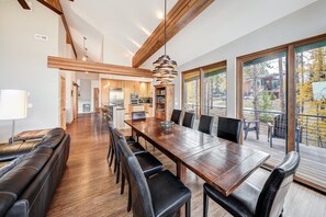 A spacious dining area with a long wooden table and black leather chairs, a kitchen in the background, and a balcony with a view of trees. - NEW Cloud 9 Cabin in Warrior's Mark near Peak 9 & 10. Amazing Views & Mtn Access! (Breckenridge)