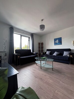 Een moderne woonkamer met een donkere bank, een glazen salontafel en een groot raam met gordijnen.
