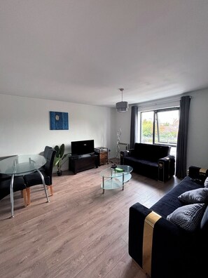 A modern living room with a glass coffee table, a black sofa, a glass dining table with chairs, and a television. - 2BR Manchester Apartment near Old Trafford (Manchester)